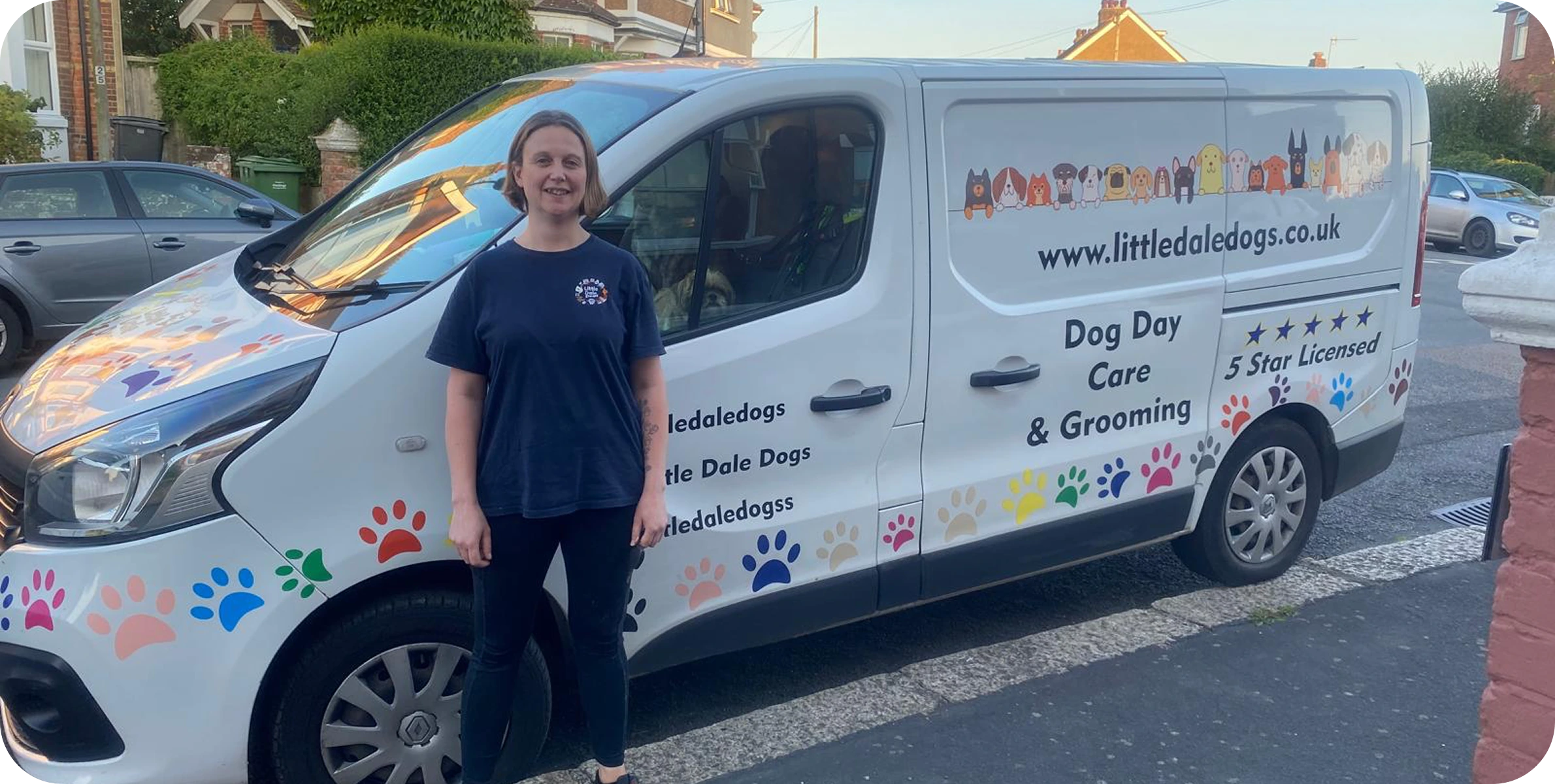 A woman standing next to a white van with colorful paw prints and the logo for a dog care and grooming service. The van is parked on a residential street during the day.